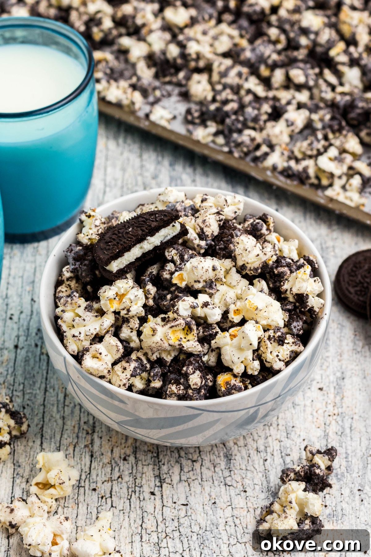 A bowl of freshly made Oreo popcorn, featuring fluffy popcorn, crushed Oreo cookies, and a delicate drizzle of white chocolate, with blue glasses of milk in the background. 