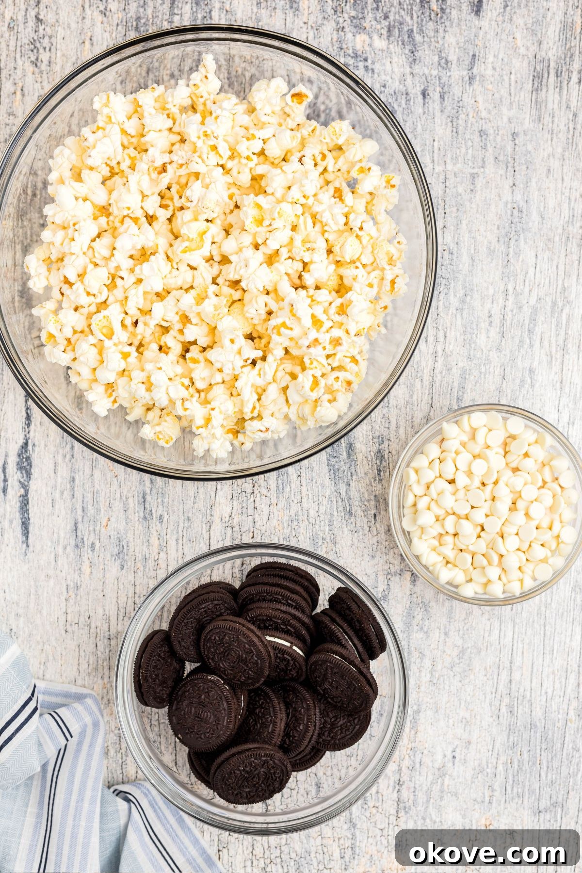 Ingredients for making Oreo popcorn: popped popcorn, Oreo cookies, and white chocolate chips, all neatly measured into clear glass bowls.