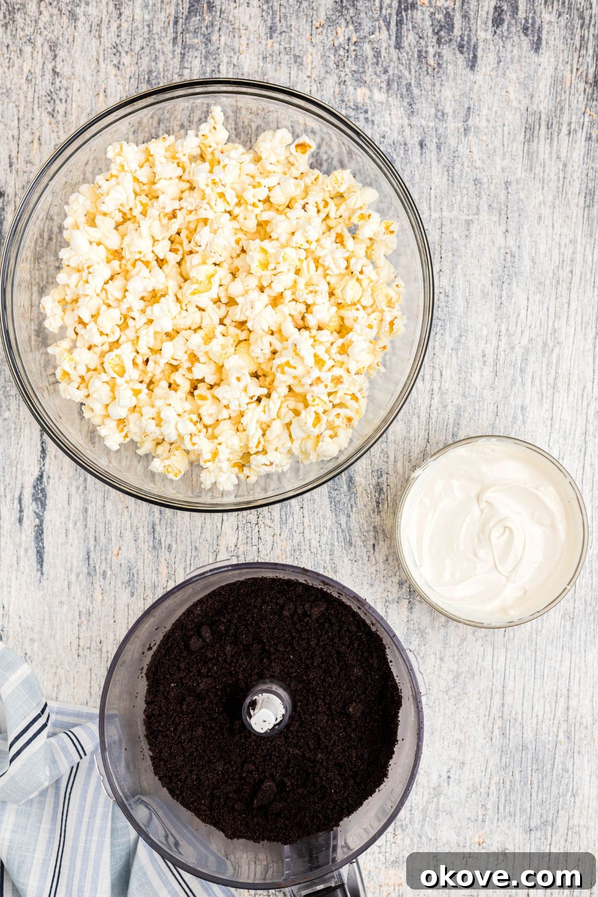 Crushed Oreo cookies in a food processor, alongside a bowl of melted white chocolate and a bowl of popcorn, on a rustic blue wooden table.