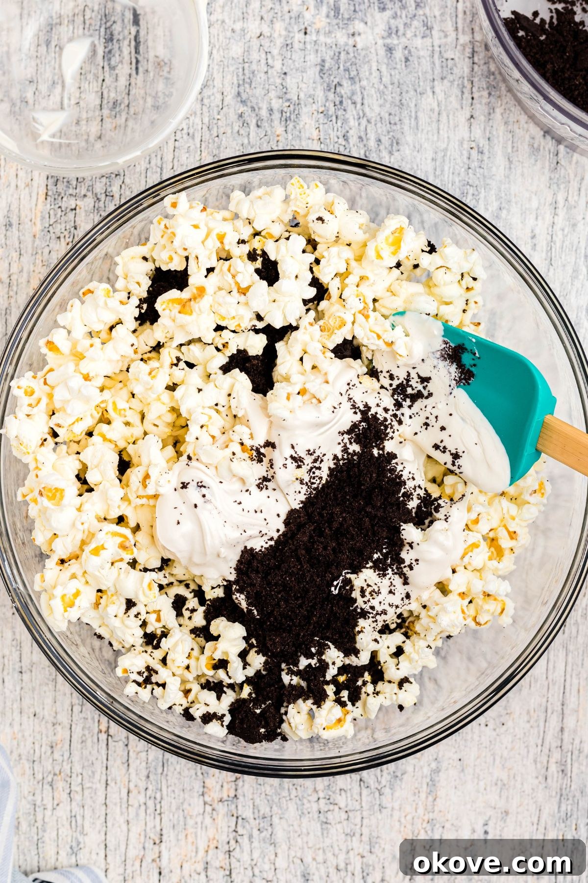 Melted white chocolate being gently mixed with popcorn and Oreo crumbs in a clear glass bowl using a spatula.