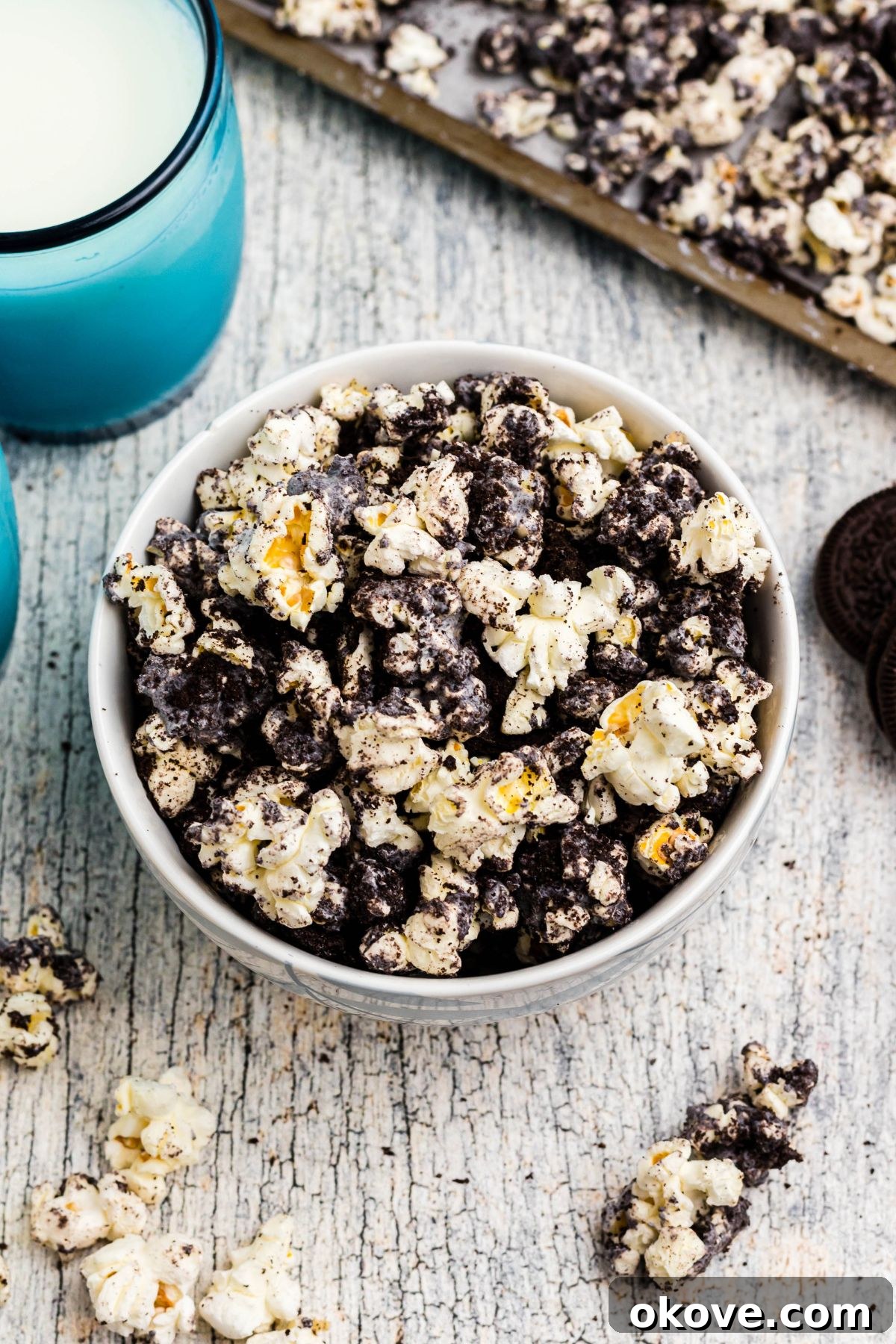 A large bowl overflowing with delicious Oreo popcorn, set on a table alongside blue glasses of milk, with scattered popcorn pieces adding to the inviting scene.