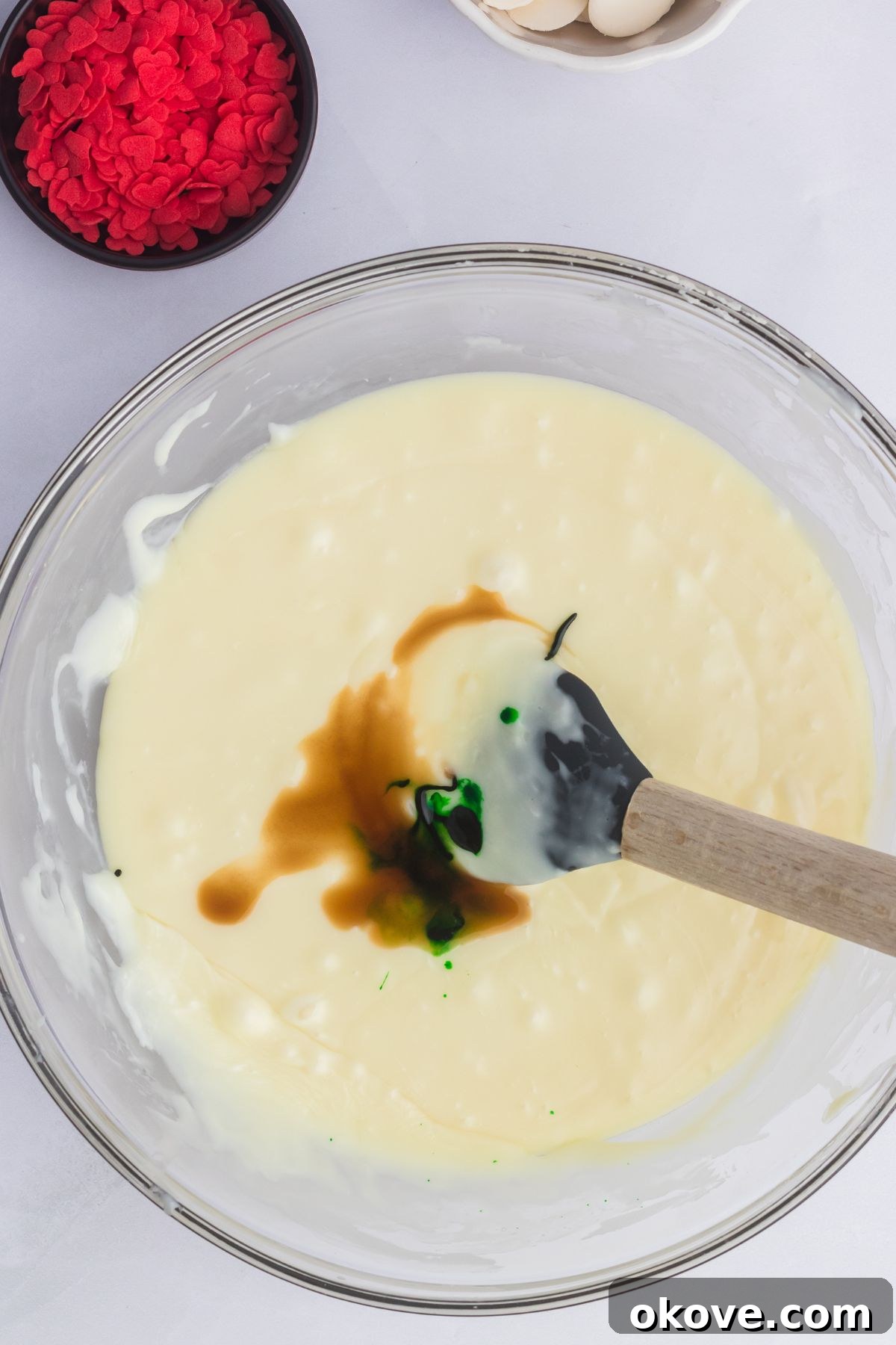 Grinch's Heart-Melting Fudge 5 Melted white chocolate with green food coloring being added to it in a large clear glass bowl. The color is starting to spread.