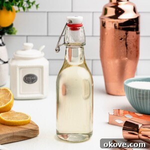 Clear glass bottle of simple syrup on a table, with lemon slices and bar mixing tools next to the bottle.