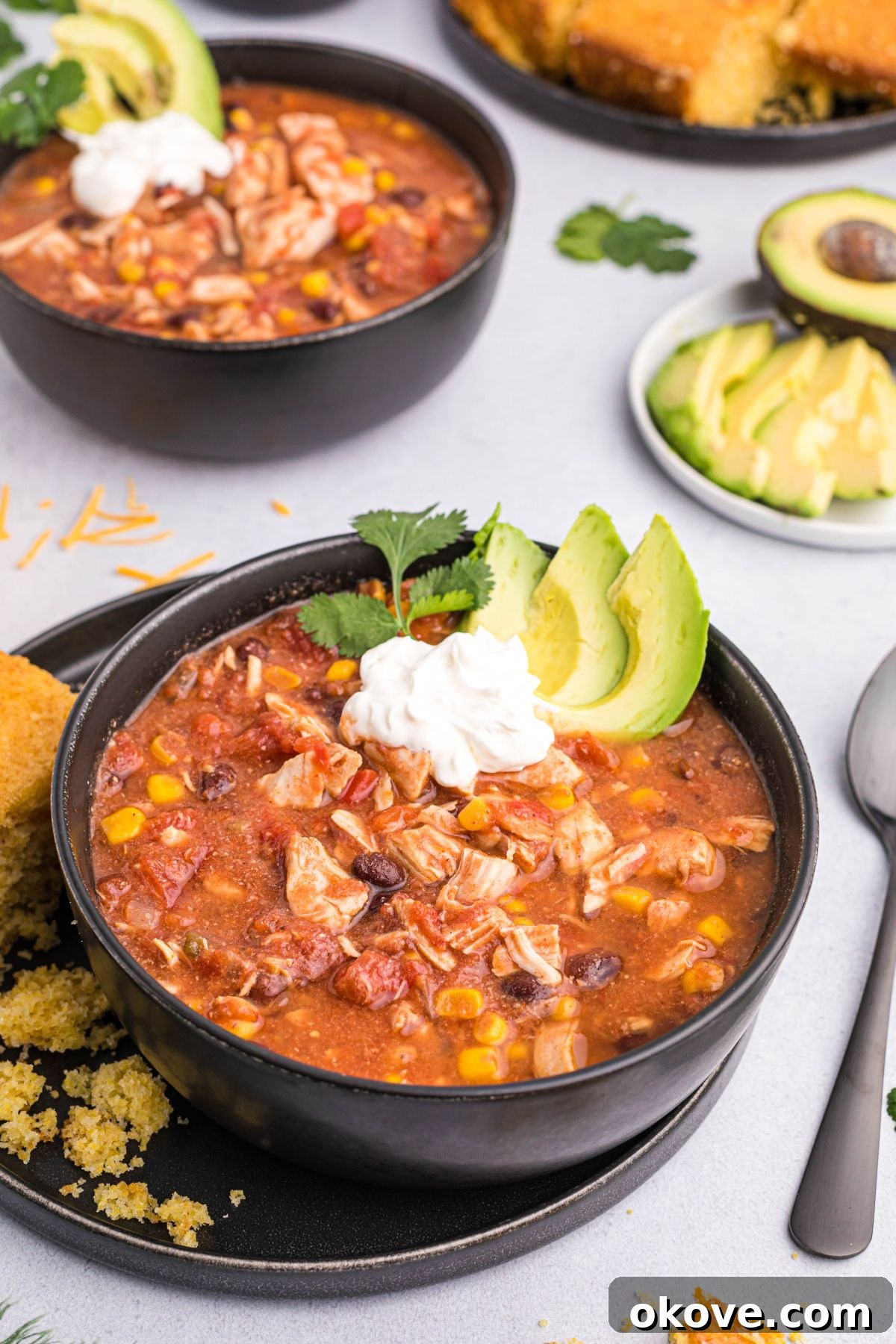 Slow cooker chicken chili in a black bowl topped with fresh avocado slices and creamy sour cream. A spoon is ready to serve.