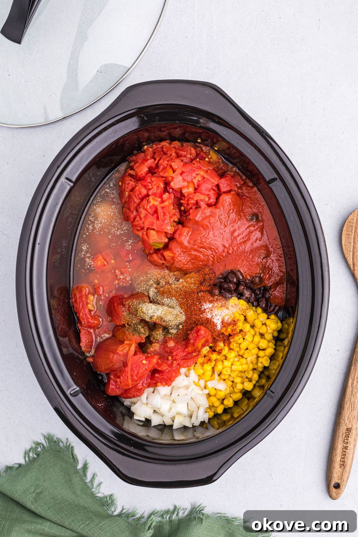 Various chili ingredients including tomatoes, onion, corn, broth, and seasonings layered over chicken in a slow cooker bowl.