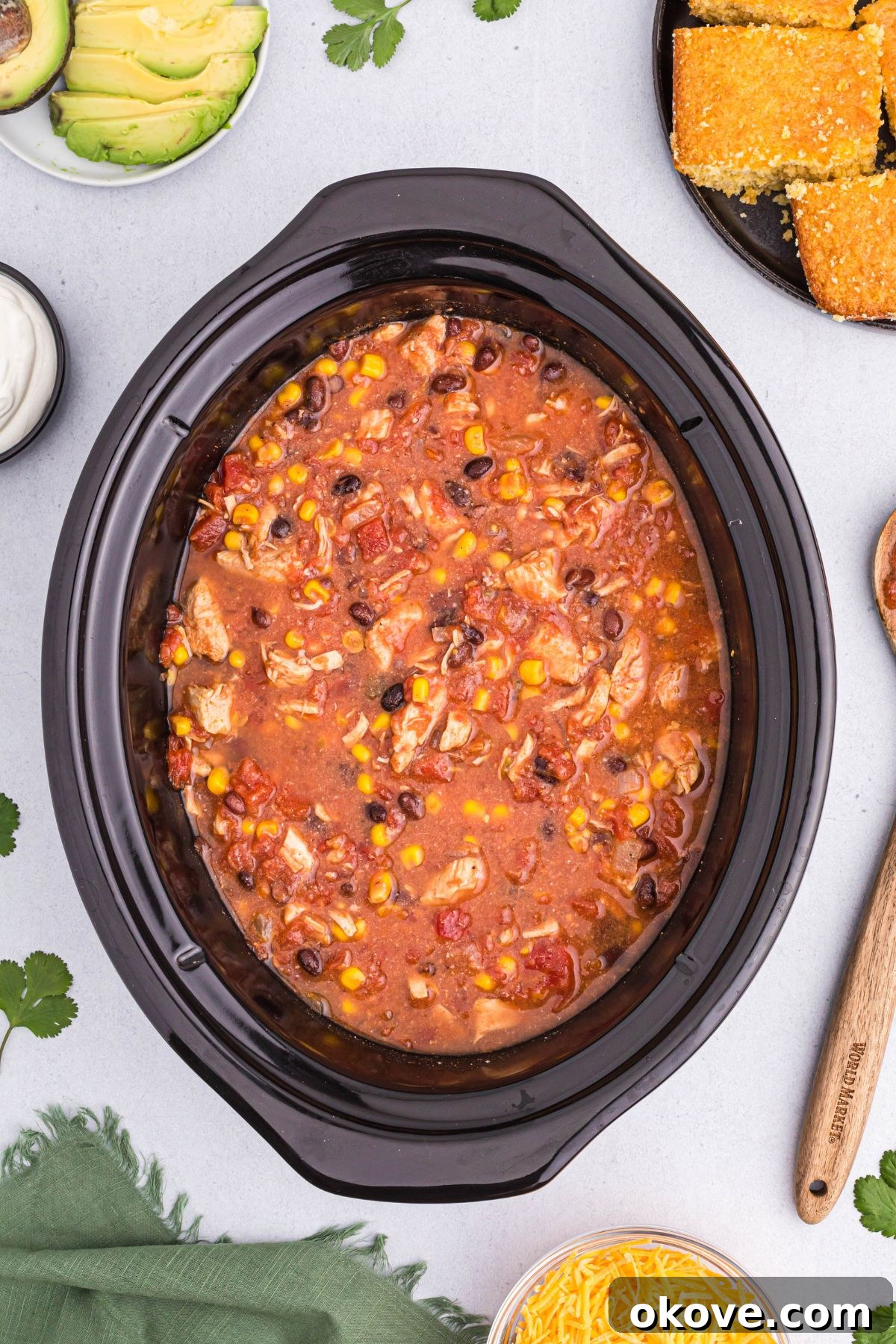 A hearty bowl of cooked chili fresh from the slow cooker, ready to be served with cornbread and various toppings laid out on the table.