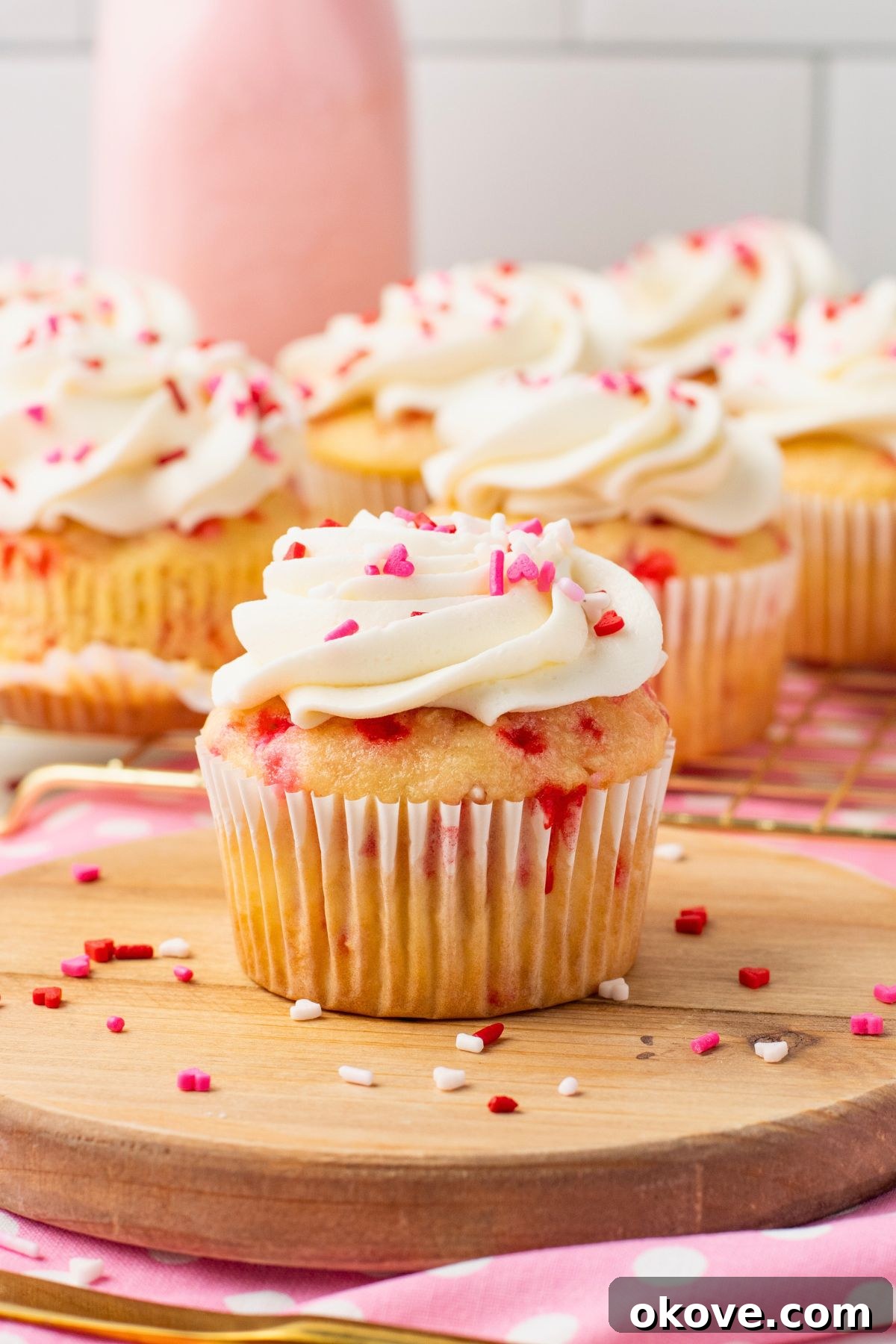 Blushing Sweet Treats 2 Vanilla cupcakes filled with pink sprinkles topped with white frosting and pink sprinkles with pink milk in the background.
