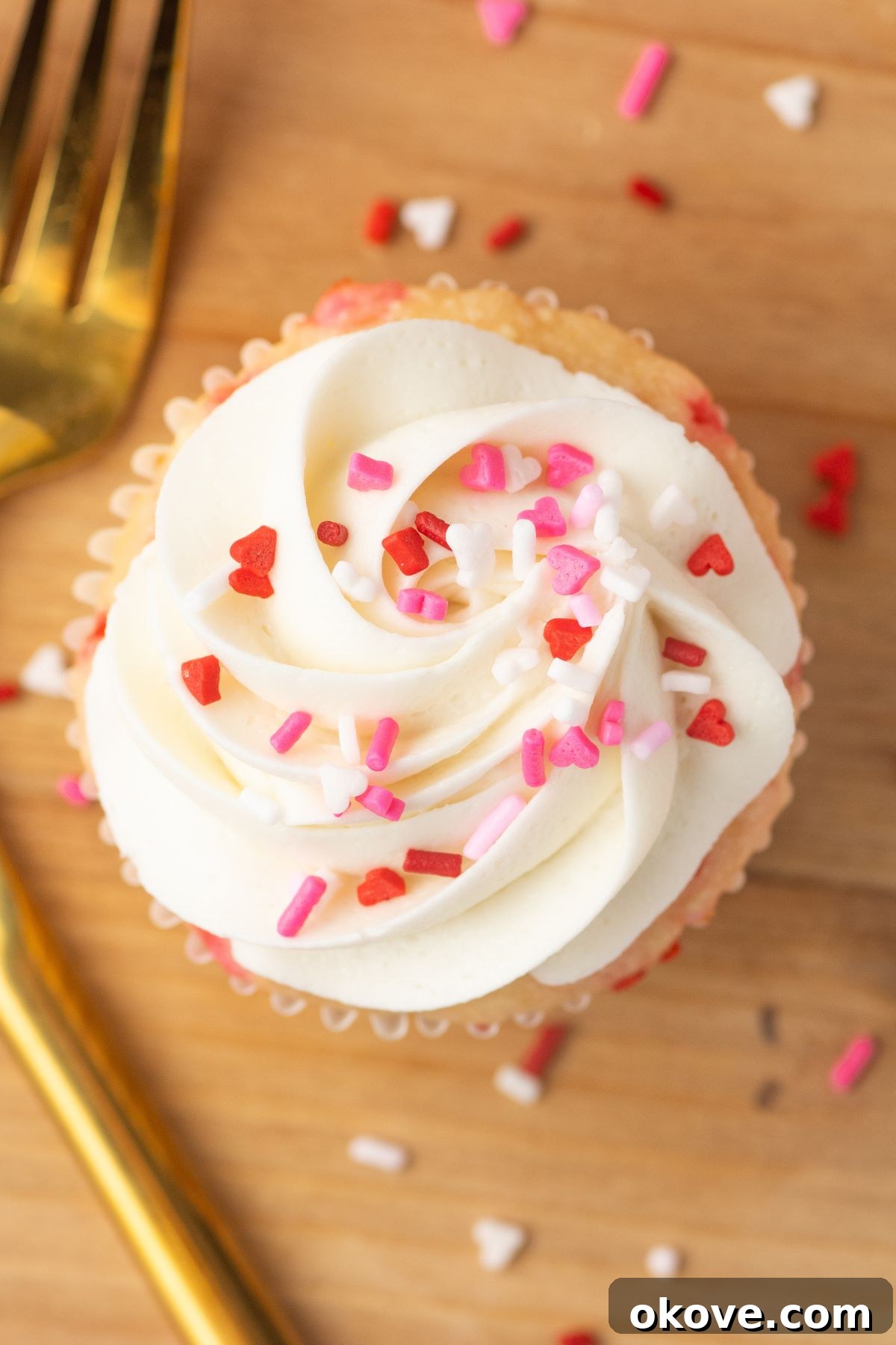 Blushing Sweet Treats 11 White cupcakes with white frosting and pink sprinkles on a wooden cutting board.