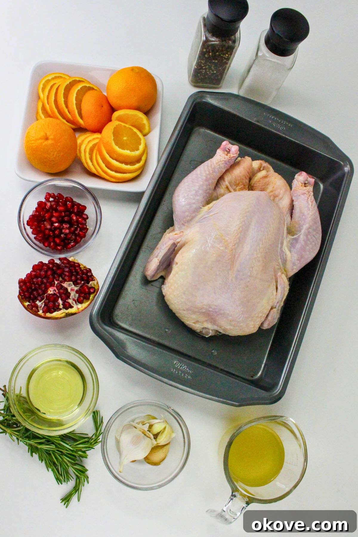 An uncooked whole chicken with fresh oranges, rosemary, and garlic cloves measured out on a pristine marble table, ready for preparation.