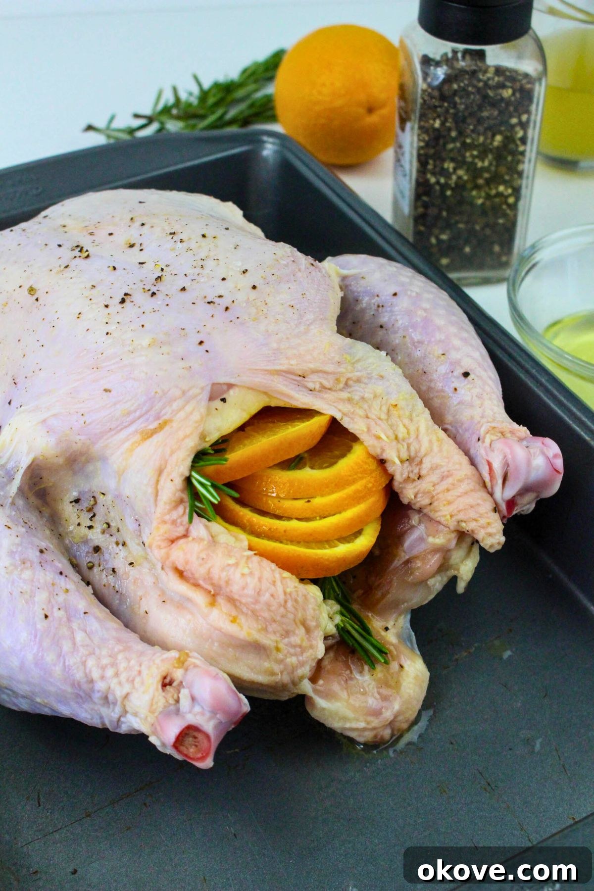 Uncooked whole chicken stuffed with fresh orange slices, rosemary sprigs, and smashed garlic cloves, nestled in a roasting pan lined with foil.
