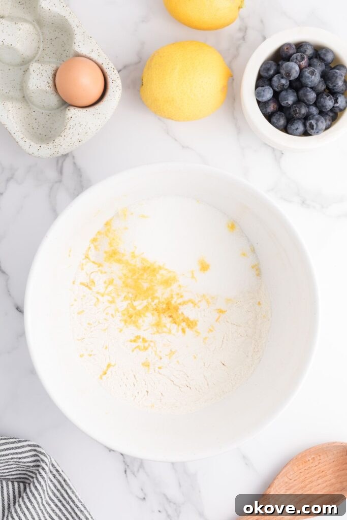 Dry ingredients like flour, sugar, salt, and lemon zest being mixed in a large white bowl with a whisk, ensuring an even distribution of flavors.