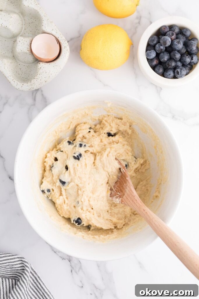 Fresh blueberries being gently mixed into a thick, pale yellow muffin batter in a white bowl with a wooden spoon. The batter is studded with bright blueberries and specks of lemon zest.