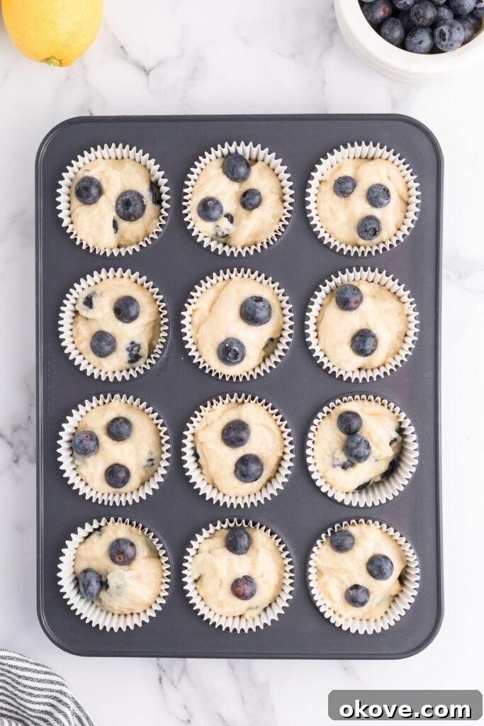 Unbaked muffin batter, generously flecked with fresh blueberries and lemon zest, neatly scooped into white paper cupcake liners arranged in a muffin tin, patiently awaiting its turn in the oven.