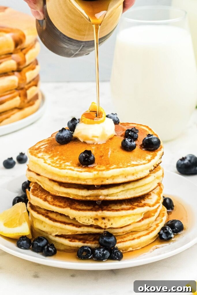 Golden fluffy lemon pancakes stacked on a white plate with syrup being poured on top. 