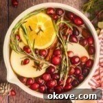 Cream colored saucepan filled with water, cranberries, fruit slices, and other ingredients to make a Christmas simmer pot on a wooden table with a red and white striped linen.