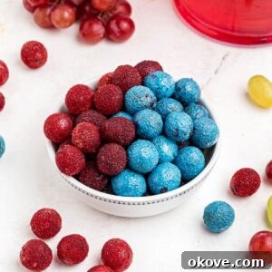 Red and blue candy grapes stacked in a white bowl with grapes scattered on a marble table.