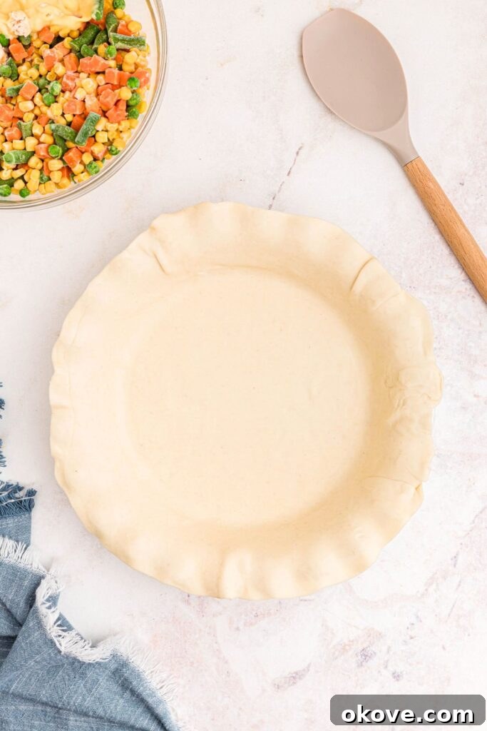 Quick and Cozy Chicken Pot Pie 4 Rolled out pie crust in a pie plate with a bowl of veggies and other filling ingredients on a marble table, ready for assembly.