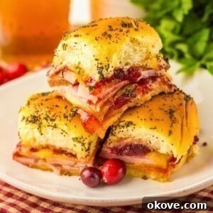 Festive Finger Sandwiches 9 Cranberry and ham sliders stacked on a white plate with red cranberries as a garnish.