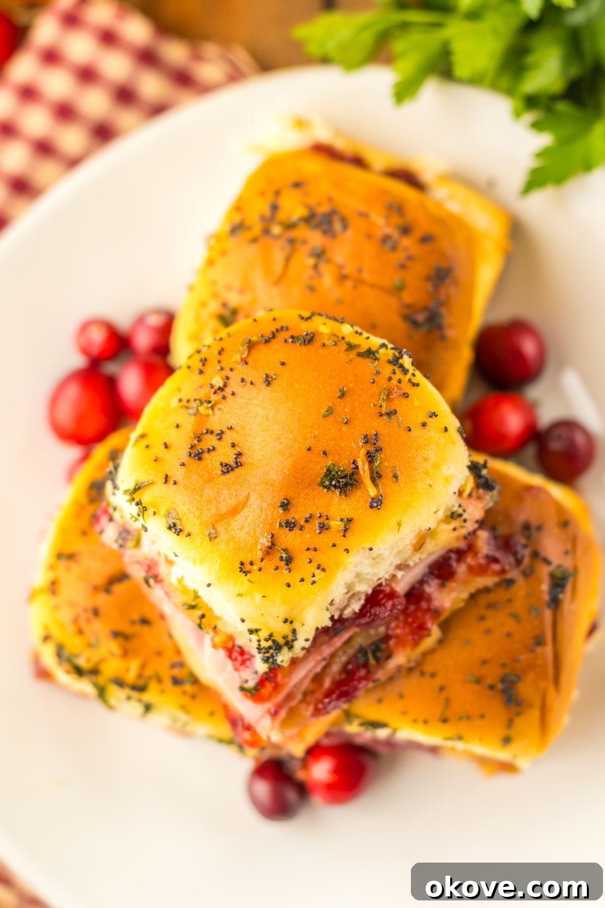 Festive Finger Sandwiches 4 Cranberry and ham sliders stacked on a white plate with red cranberries as a garnish.
