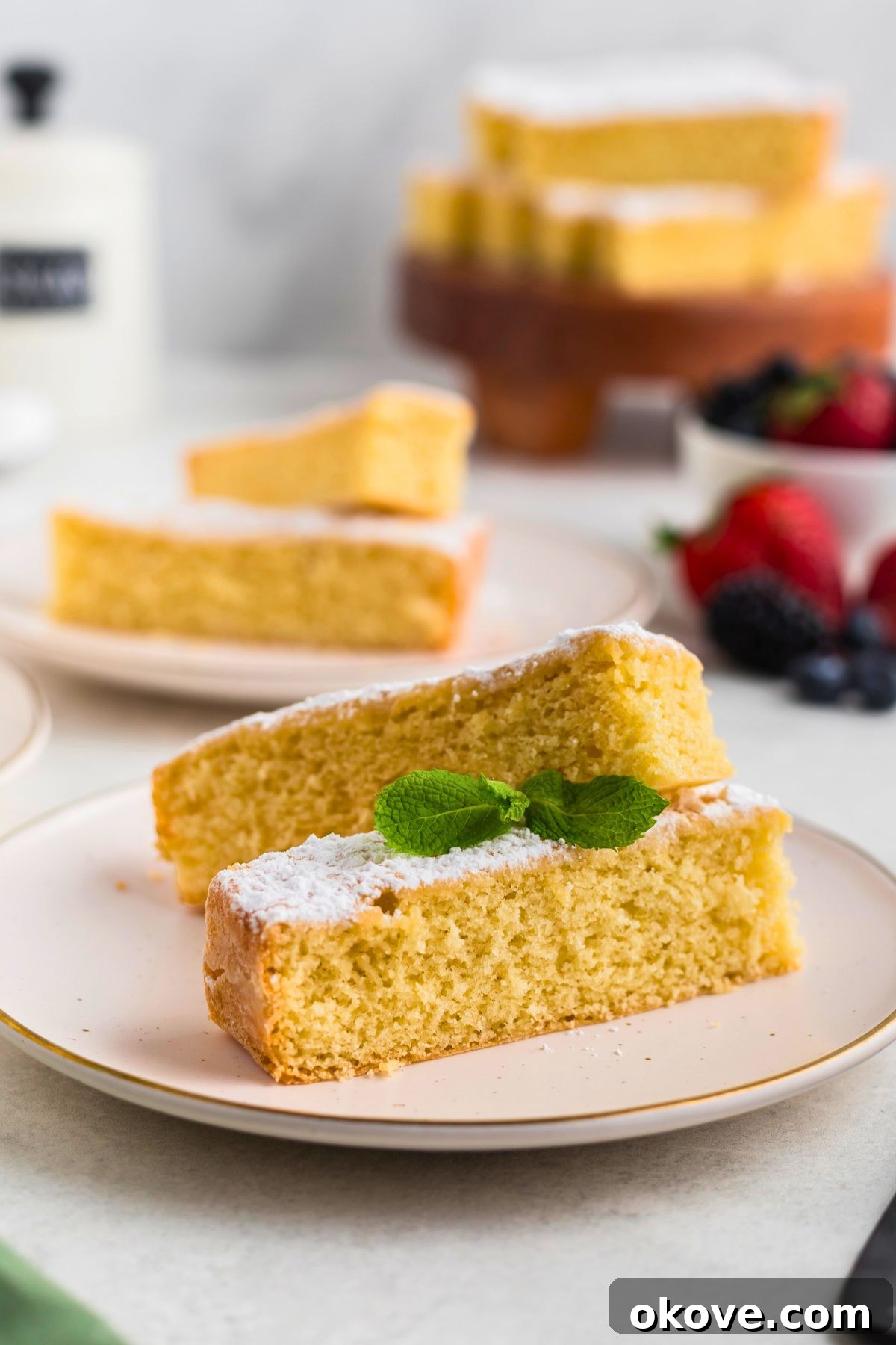 Buttery Bliss Cake 2 Golden yellow butter cake slices on white plates topped with powdered sugar with berries on the table.