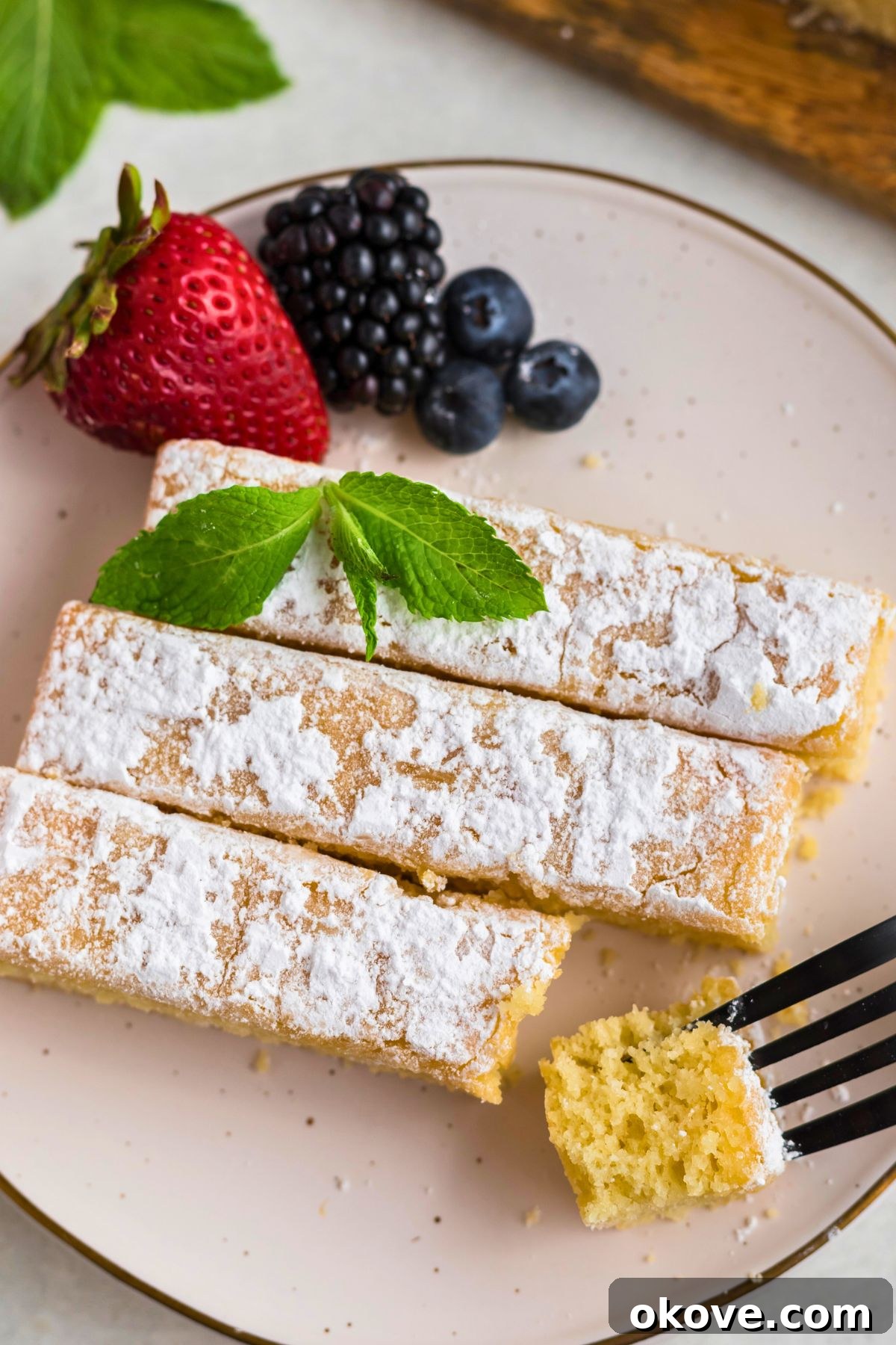 Buttery Bliss Cake 9 Golden yellow butter cake slices on white plates topped with powdered sugar with berries on the table, with a fork taking a small bite.