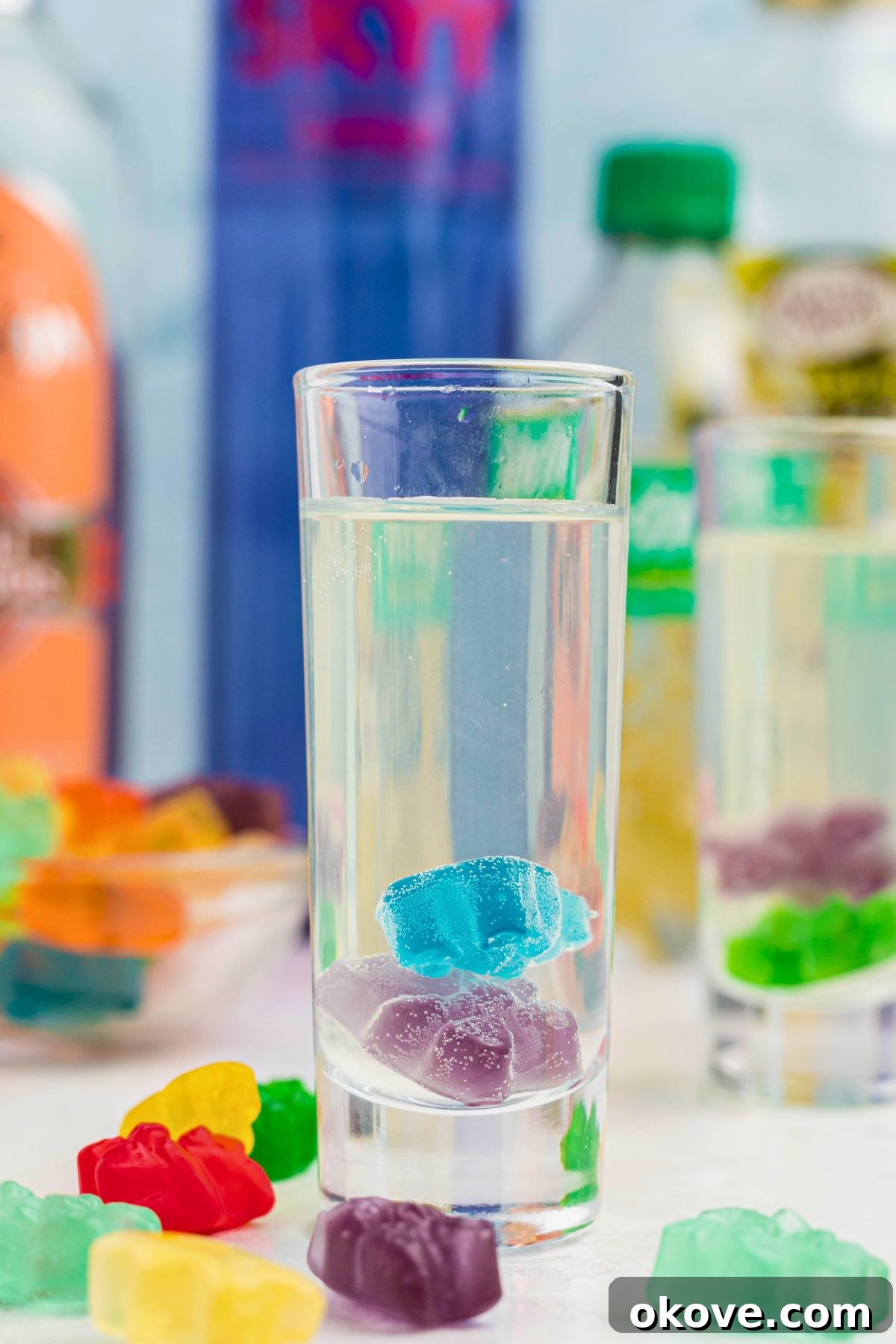 Beary Boozy Bliss 2 Purple and blue gummy bear in a clear shot glass with a vodka mixture poured over them, and with more gummy bears scattered on the table.