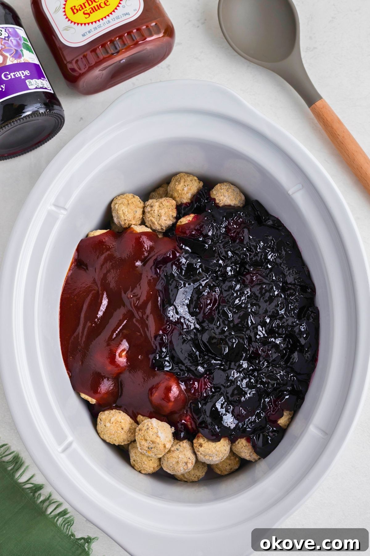 Frozen meatballs, grape jelly, and bbq sauce in a white slow cooker before cooking. Ready to be stirred and cooked.