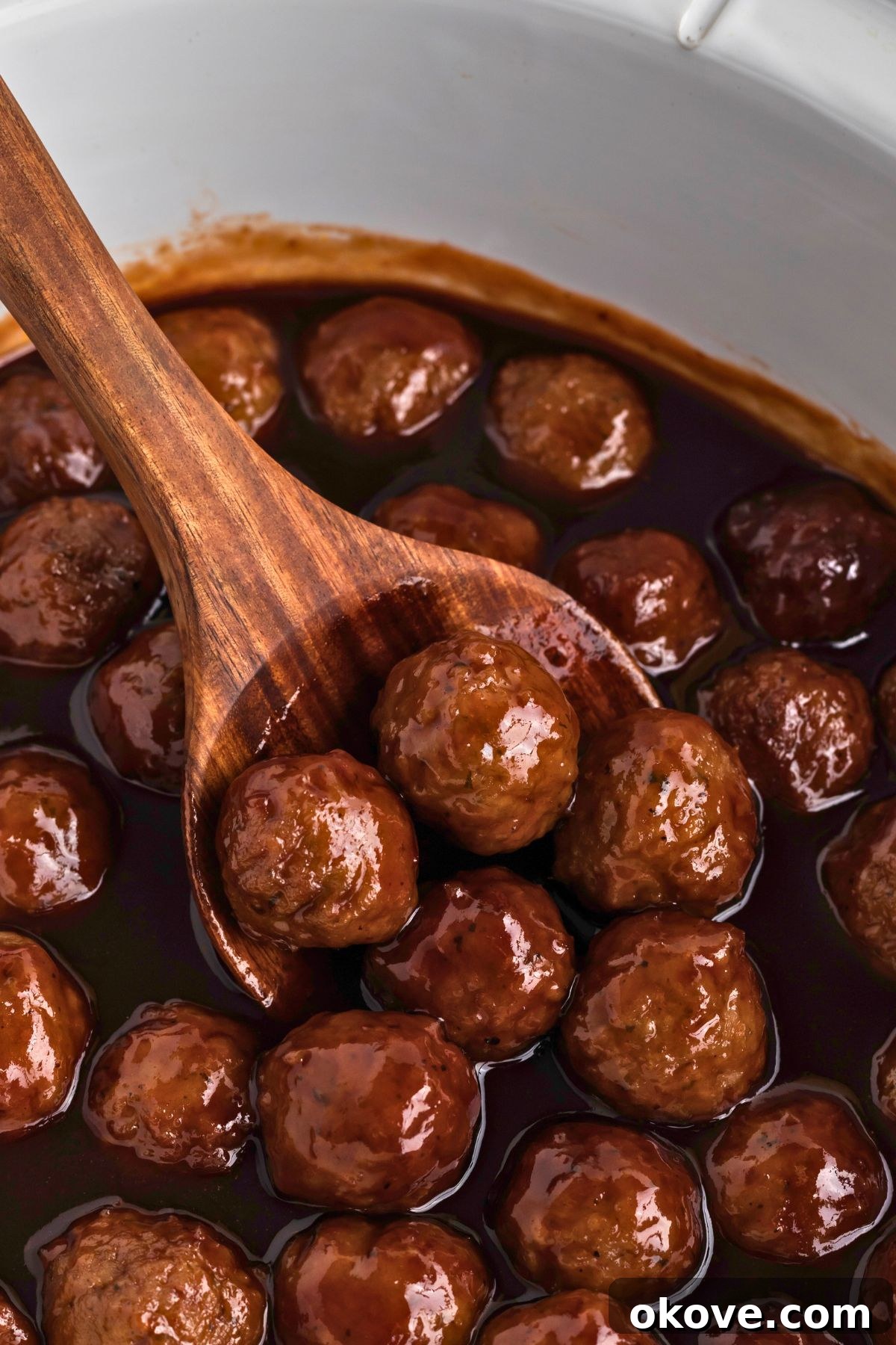 Grape jelly meatballs in a white crock pot being scooped up to serve with a wooden spoon. Ideal for parties.