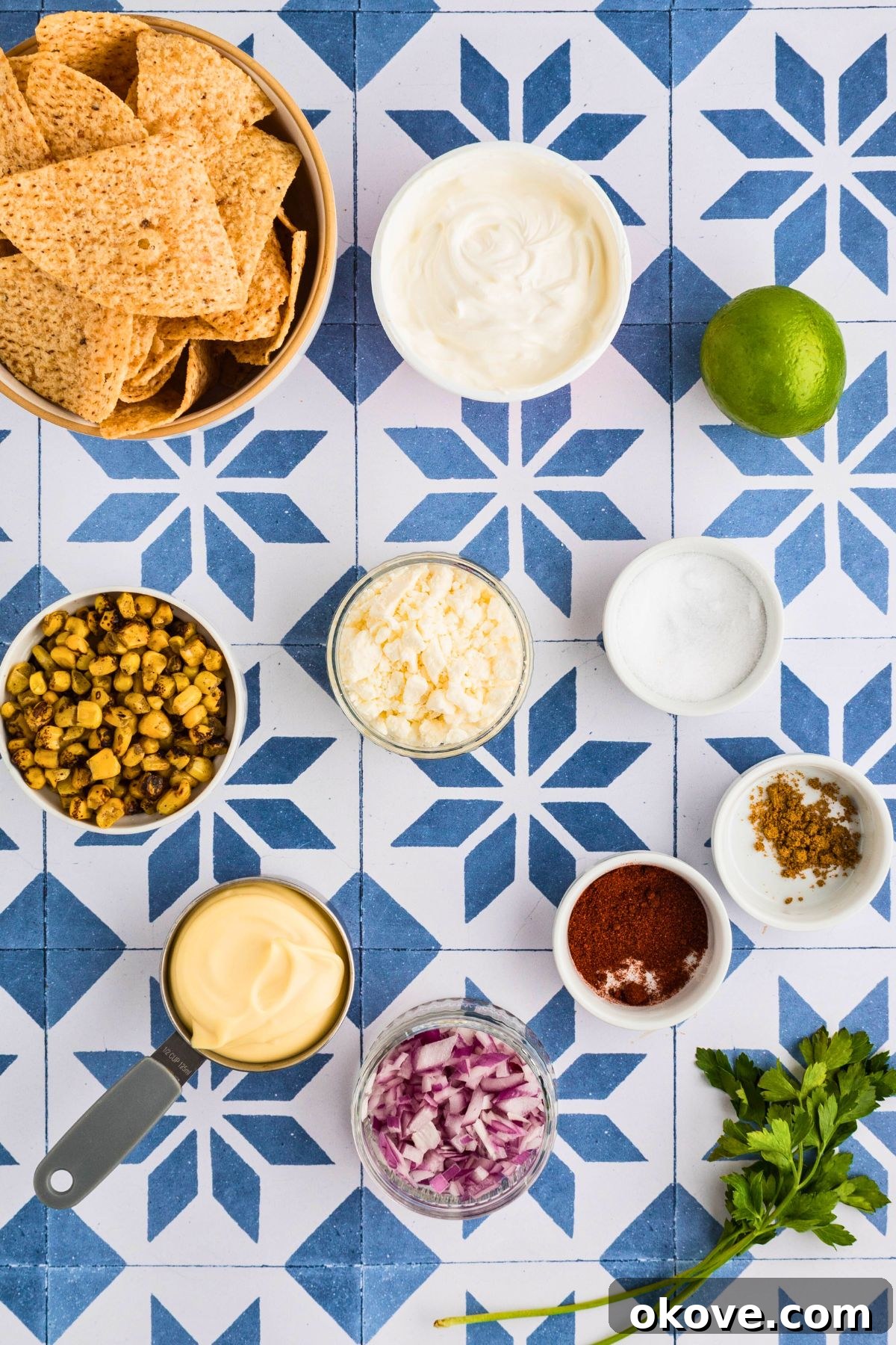 Fiesta Elote Dip 3 All the essential ingredients for Mexican Street Corn Dip, including corn, red onion, lime, and spices, measured into small bowls on a blue and white tiled table.