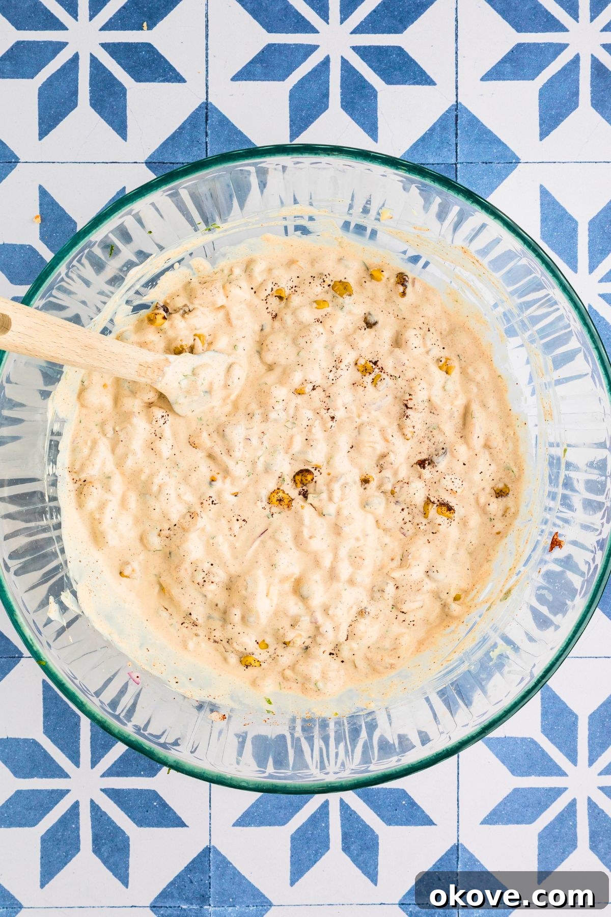 Fiesta Elote Dip 5 All Mexican street corn dip ingredients expertly mixed together in a clear glass bowl, showcasing its vibrant colors and creamy texture, resting on a blue tiled table.