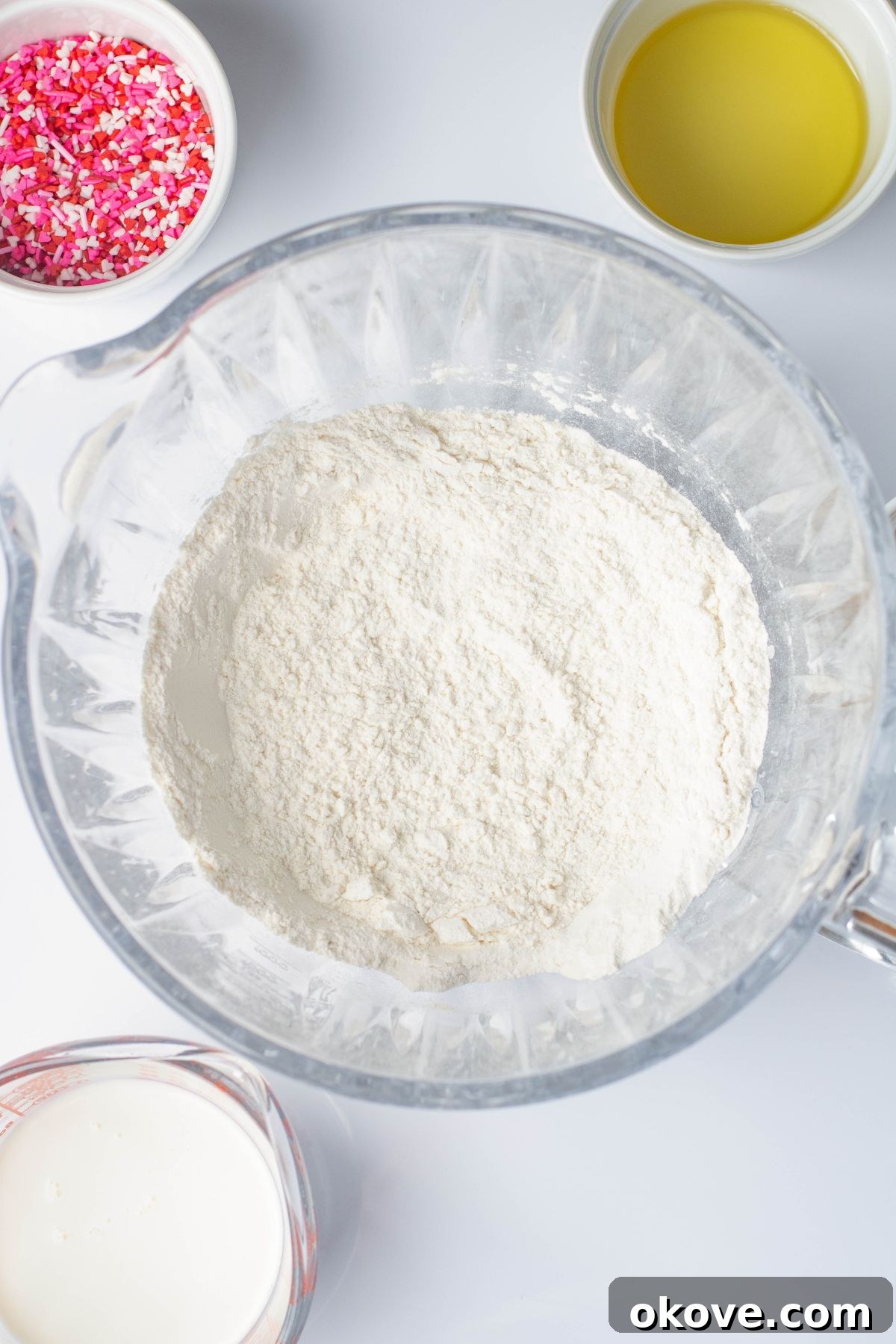 Sweetheart Pancakes 4 All the liquid and dry ingredients for pancake batter are combined in a clear mixing bowl, being gently mixed together before the addition of festive sprinkles. The batter is smooth with a few small lumps, indicating it's perfectly mixed.