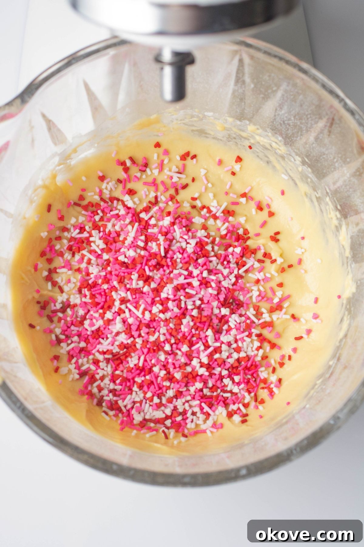 Sweetheart Pancakes 5 Vibrant Valentine's Day sprinkles are being gently mixed into a light, creamy pancake batter in a clear glass bowl, showcasing the festive colors before cooking.