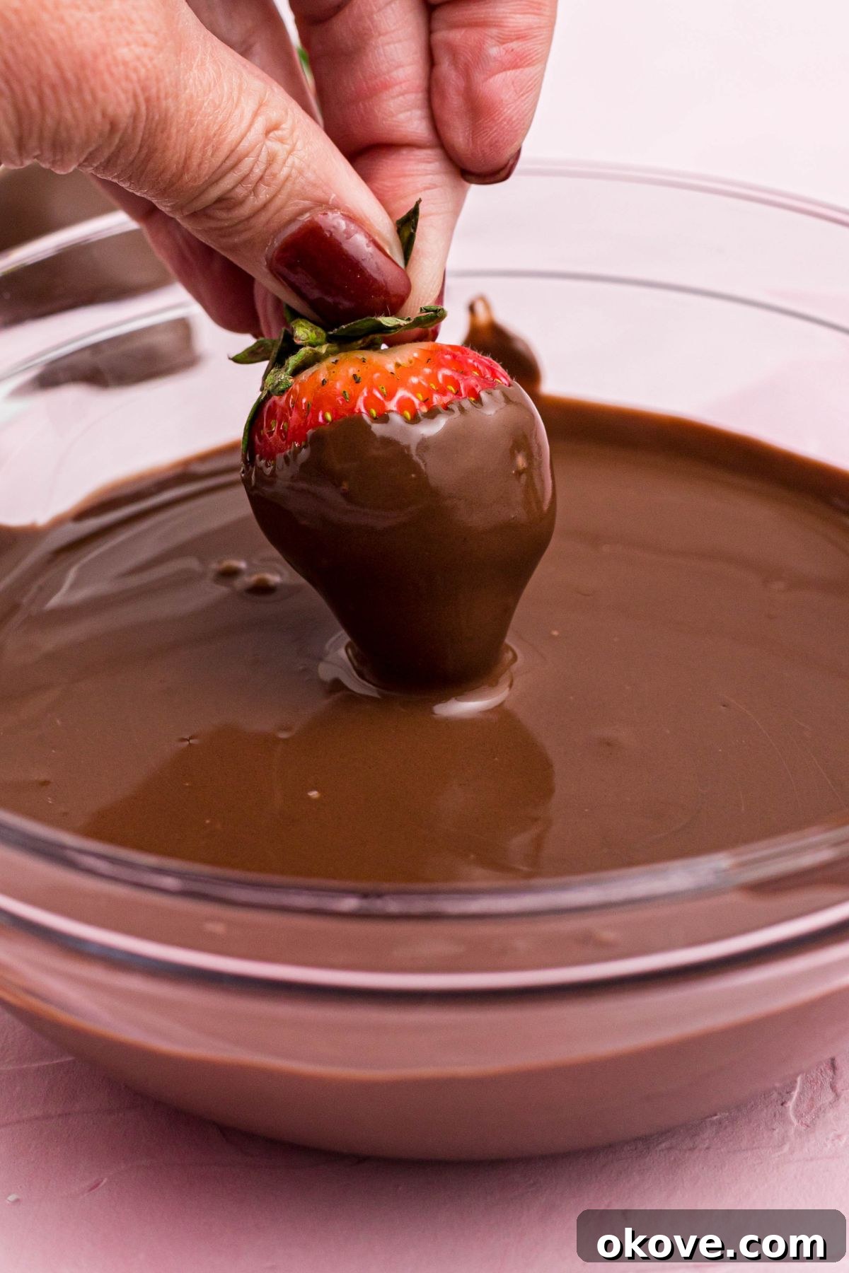 Effortless Chocolate Strawberry Delights 4 Fresh strawberry being dipped into melted milk chocolate in a clear glass bowl.