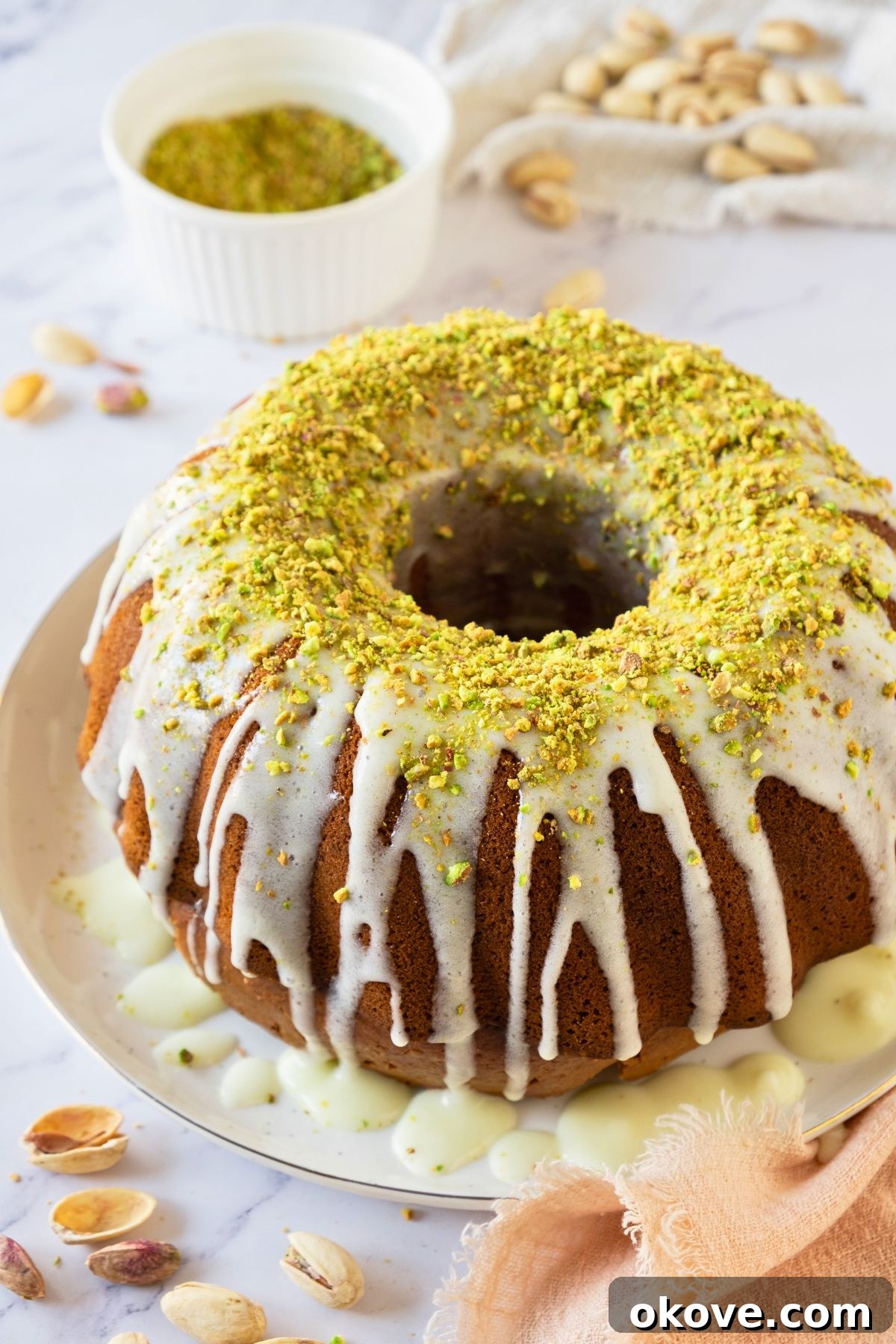 Emerald Pistachio Bundt 2 Pistachio Bundt cake on a plate with a bowl of chopped pistachios behind it on a marble table.