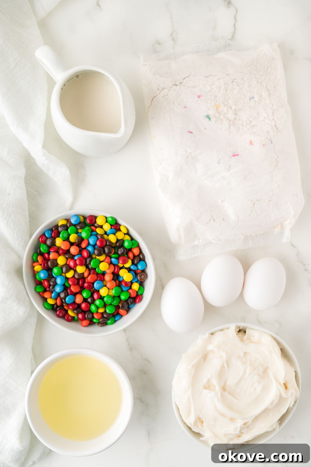 Measured ingredients for M&M cupcakes, including cake mix, eggs, milk, oil, frosting, and mini M&Ms, neatly arranged in small white bowls on a marble counter.