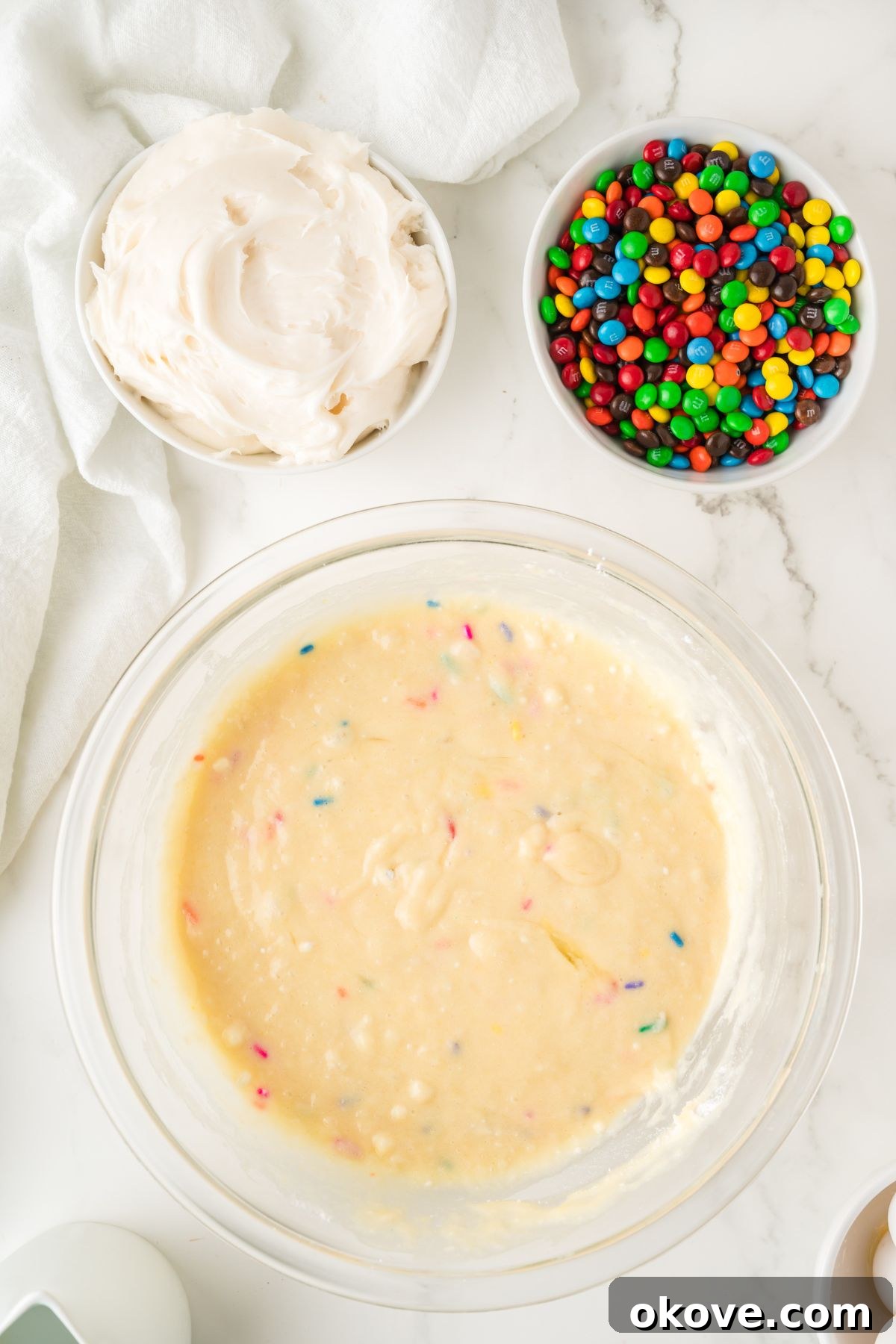 Smooth cupcake batter in a large glass bowl, with a tub of frosting and a bowl of mini M&Ms nearby on a marble surface, ready for the next step.