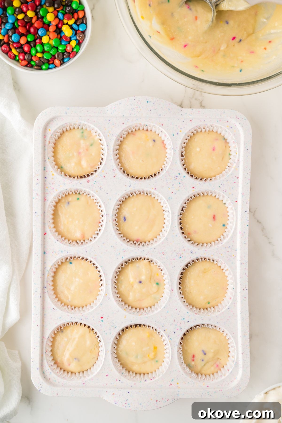 Cupcake batter poured into a muffin pan, with a bowl of colorful mini M&Ms nearby, ready to be baked.