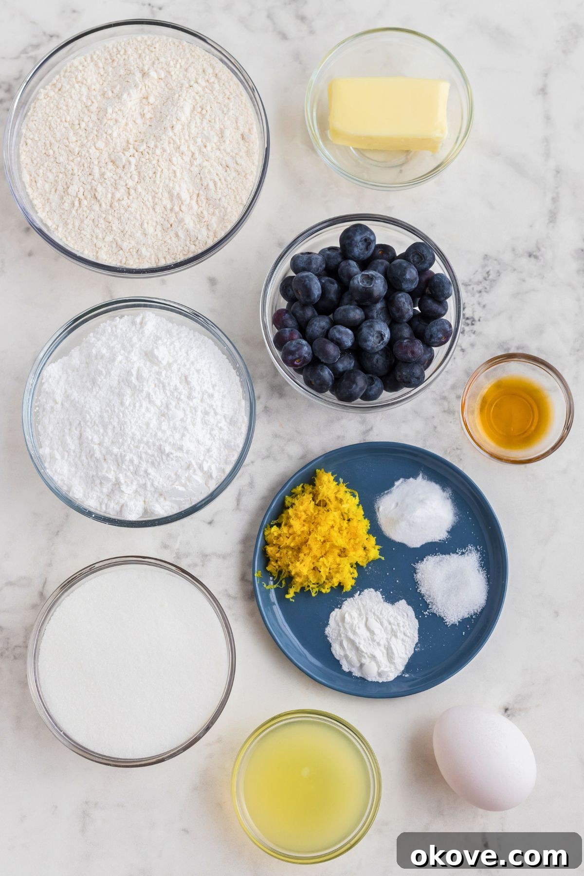 Tangy Blueberry Lemon Delights 3 Carefully measured ingredients for lemon blueberry cookies, including flour, sugar, butter, lemons, and blueberries, arranged in small bowls on a elegant marble countertop.