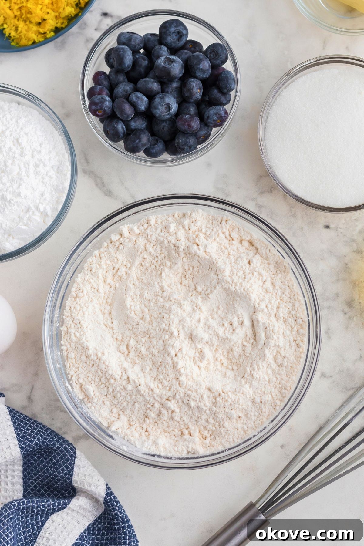 Tangy Blueberry Lemon Delights 4 Dry ingredients for cookies, including flour, baking powder, baking soda, and salt, being whisked together in glass bowls on a marble table, ready for combining.