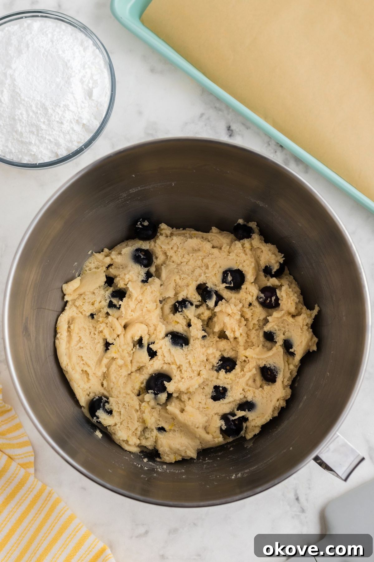 Tangy Blueberry Lemon Delights 7 A bowl of creamy cookie dough gently mixed with fresh blueberries, beside a parchment-lined baking sheet, ready for scooping and baking.