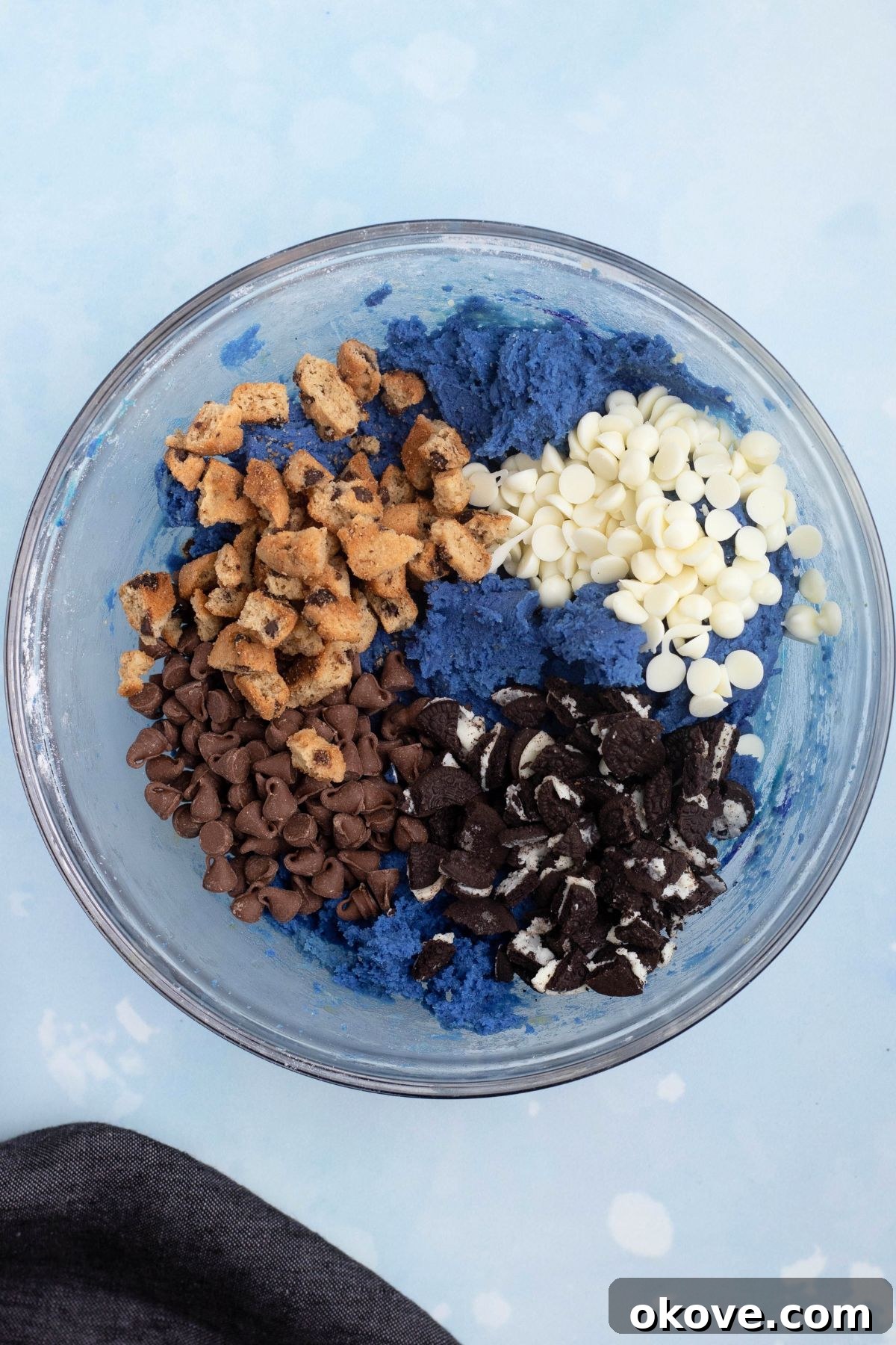 Blue Monster Munchies 6 Chopped cookies and chips being mixed in with cookie dough in a clear glass bowl.