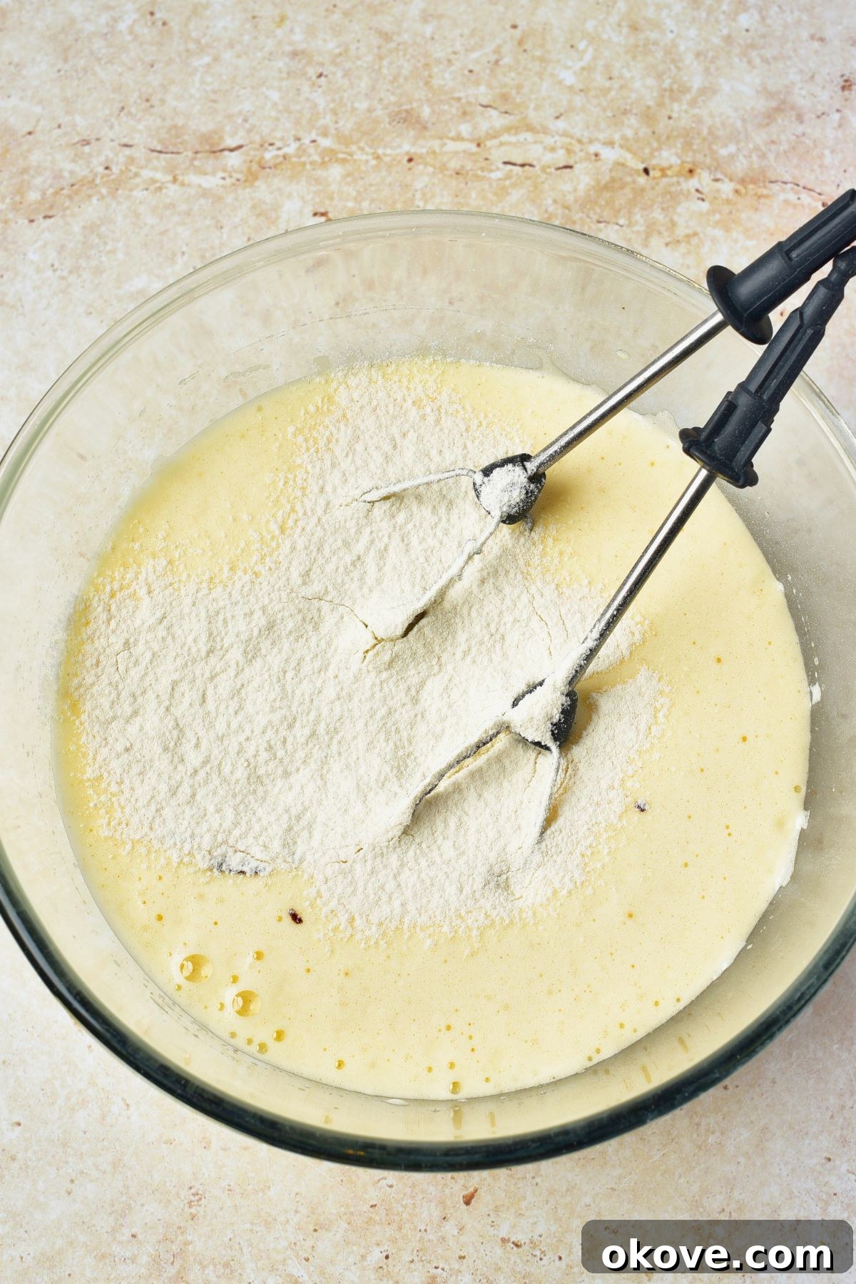 Molten Chocolate Dream 5 A creamy, pale yellow batter being vigorously mixed in a large clear glass bowl with an electric hand mixer, showing its thick and fluffy texture.