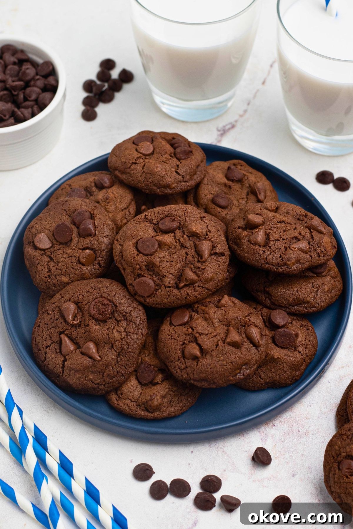 Luscious Double Chocolate Chip Cookies 2 Double chocolate chip cookies stacked on a blue plate with chocolate chips and a glass of milk on a marble table.