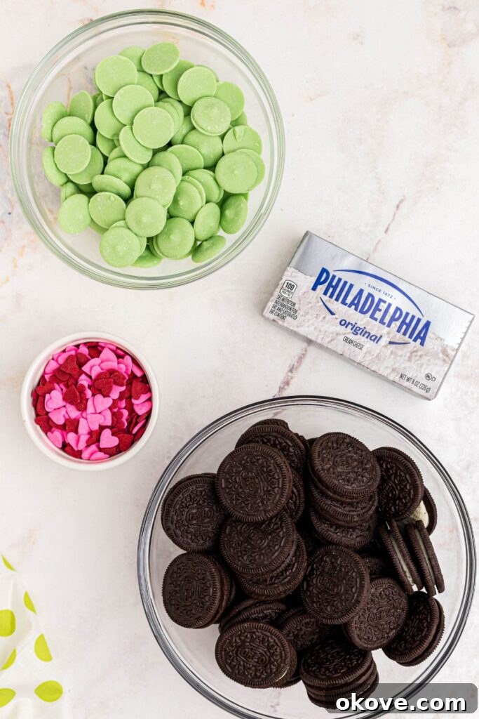 Ingredients for Grinch Oreo balls, including Oreo cookies, cream cheese, green candy melts, and red heart sprinkles, neatly arranged in small bowls on a marble surface.