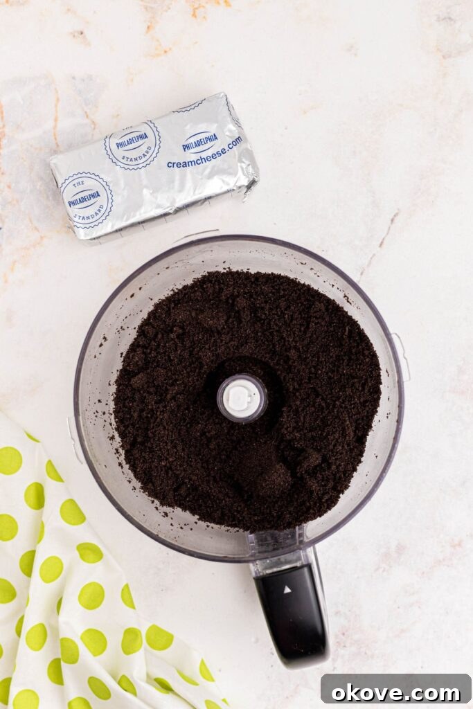 Crumbled Oreos in a food processor bowl next to a package of cream cheese on a marble counter, showing the key ingredients for the truffle base.