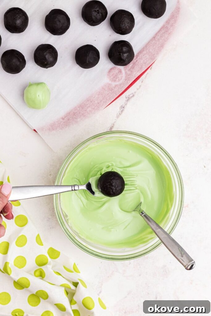 An Oreo ball being carefully dipped into a bowl of melted green candy coating with other prepared balls waiting on parchment paper.