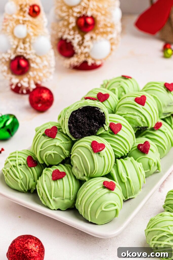 Green Grinch Oreo balls elegantly stacked on a white plate, with one truffle having a bite taken out to reveal its creamy center. Holiday ornaments decorate the surrounding table.