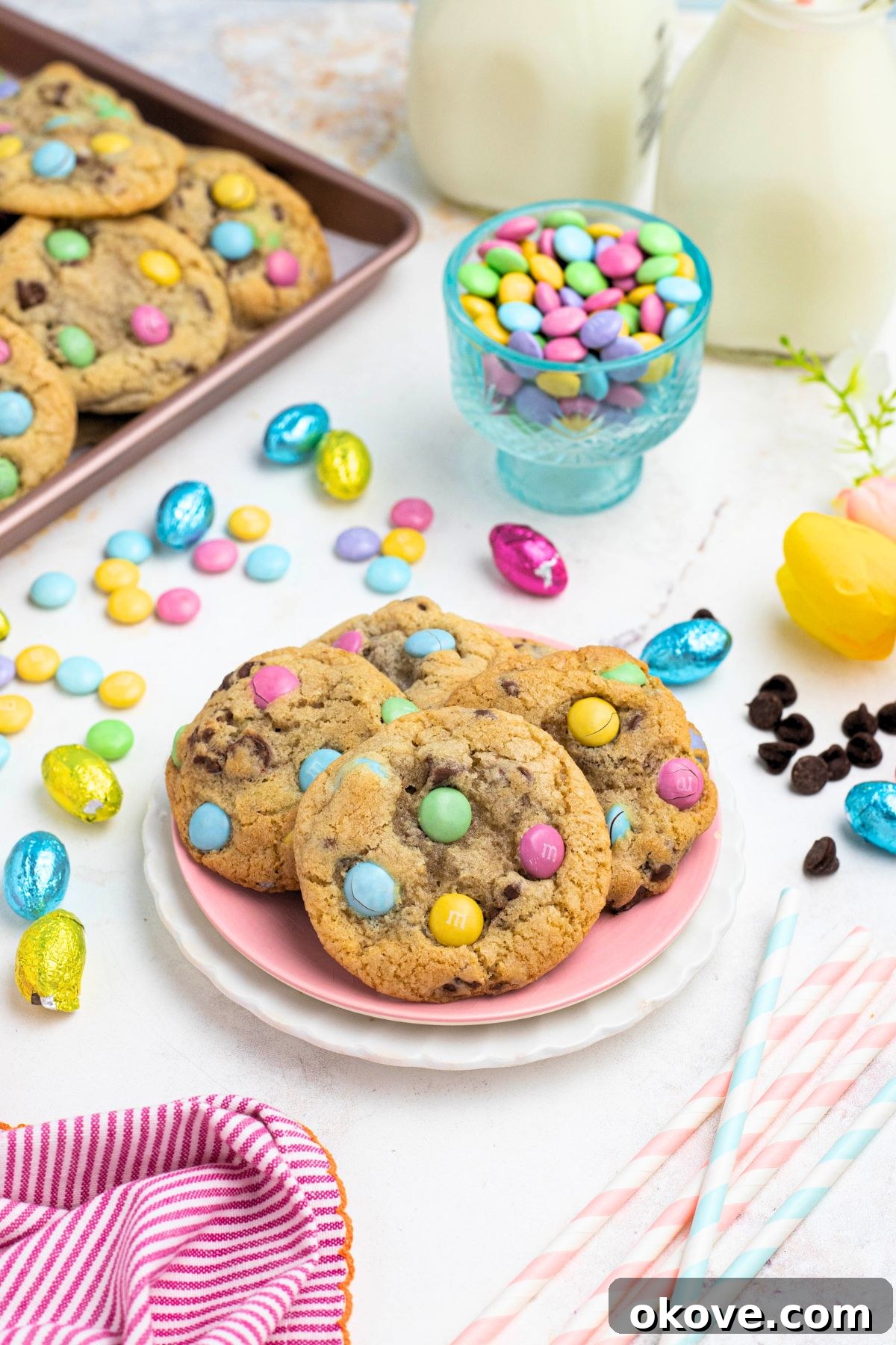 Golden chocolate chip cookies with pastel candies on plates, in front of candy dish and glasses of milk.