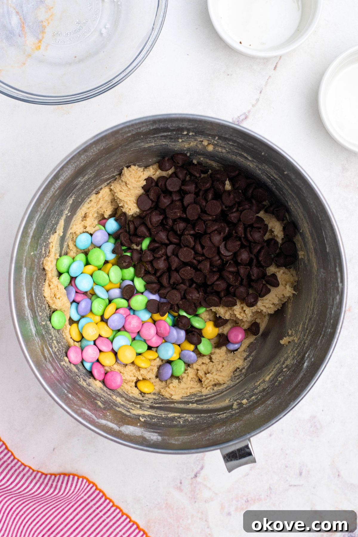 Chocolate chips, pastel m&m's and cookie dough being mixed together in a silver mixing bowl.