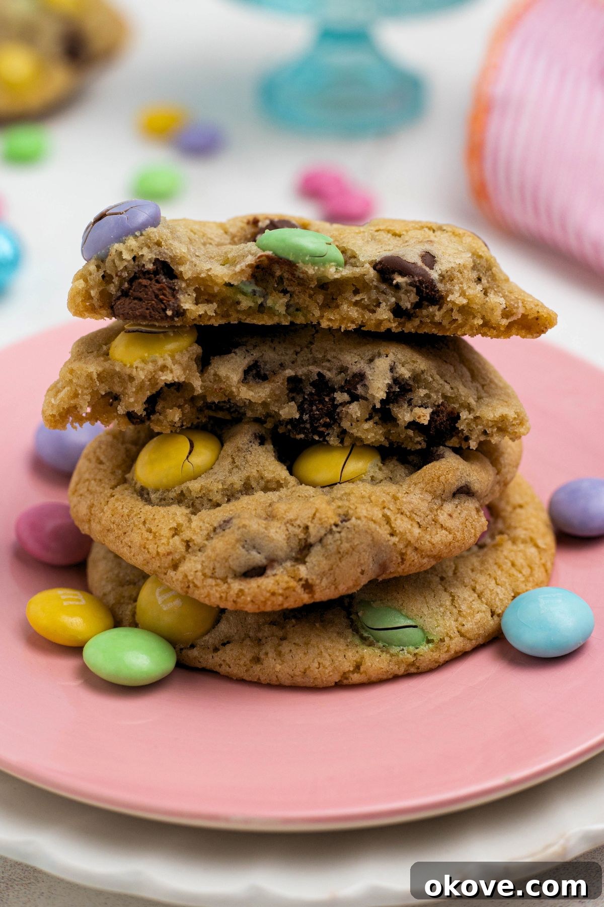 Stacked chocolate chip cookies with pastel m&m's on a pink plate with the top cookie broken in half.